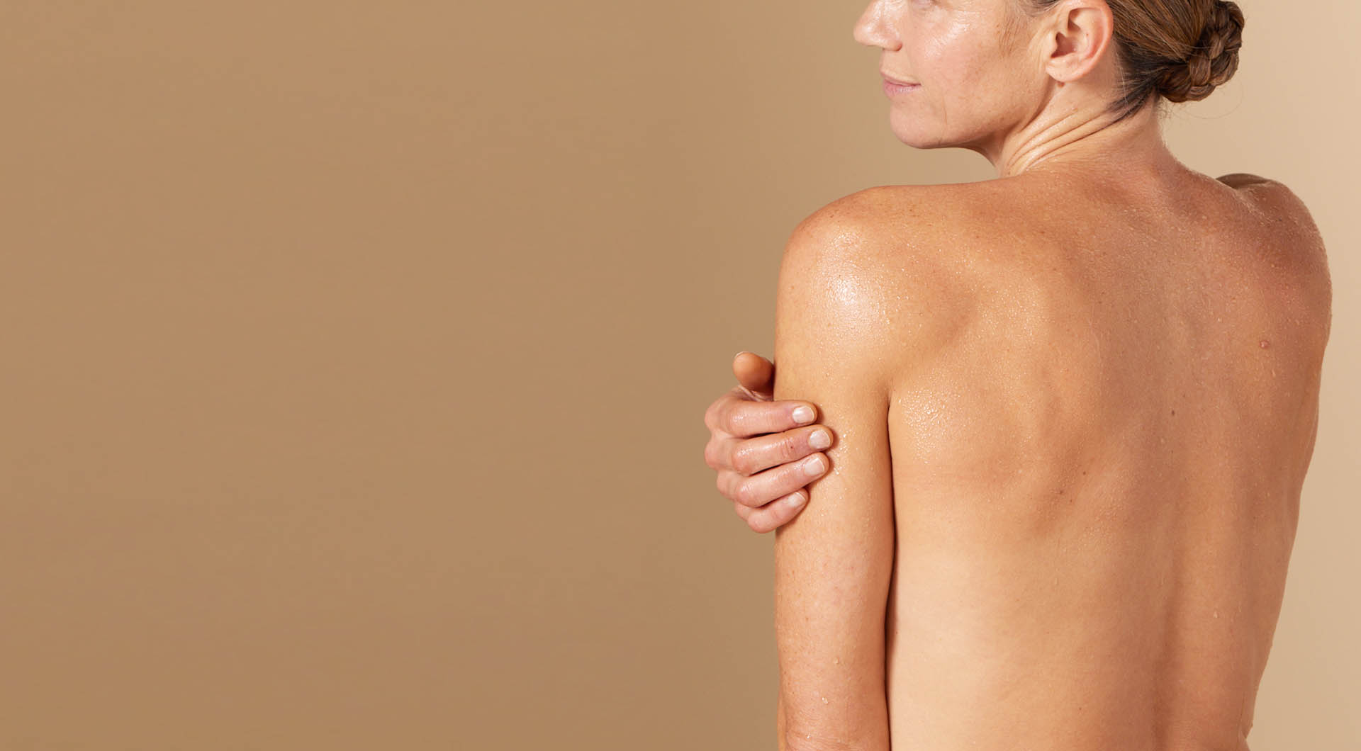 Een vrouw met een lichte huidskleur, gezien vanaf haar rug, die haar schouder inzeept met een rijk schuim tegen een beige achtergrond.